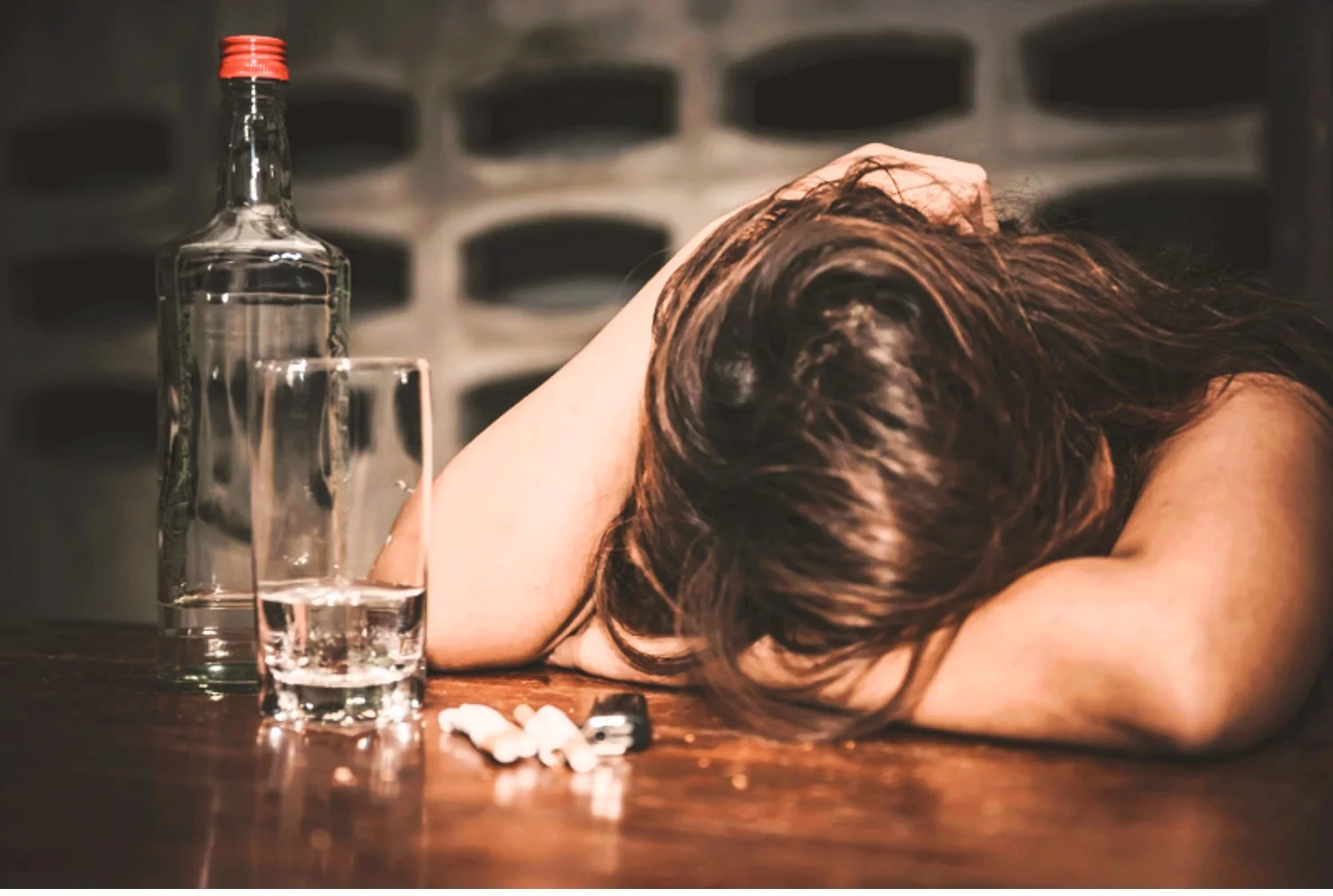 Depressed woman rests head on bar in front of an empty alcohol bottle, glass with alcohol, and pile of cigarettes
