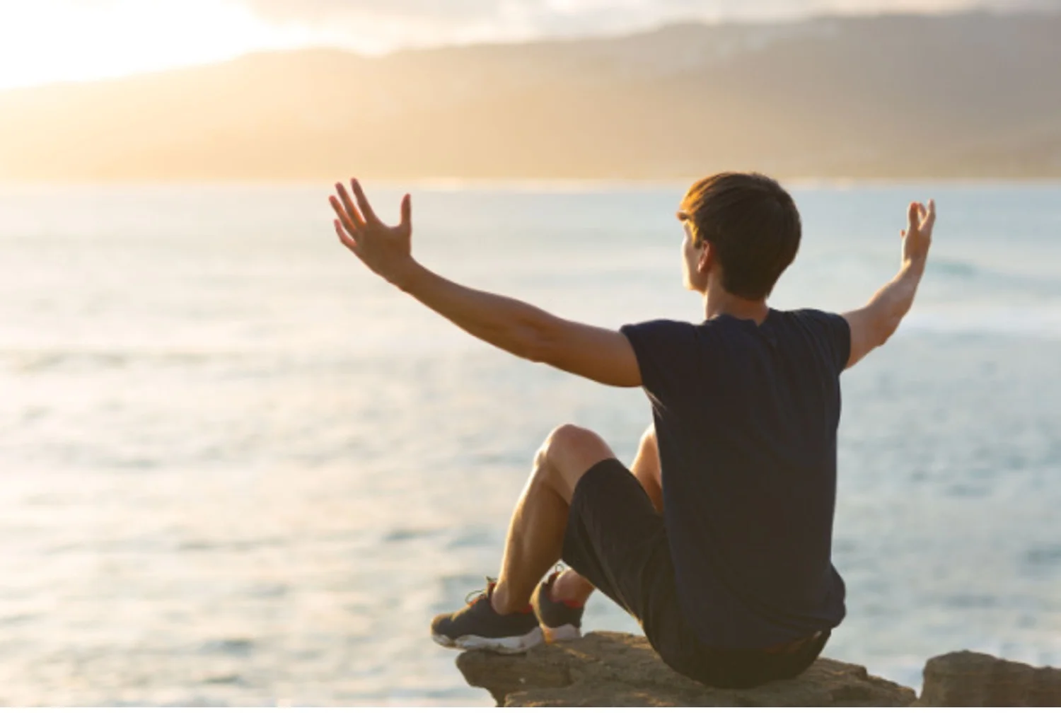 Optimistic young man sits on a rock overlooking the ocean with arms stretched out