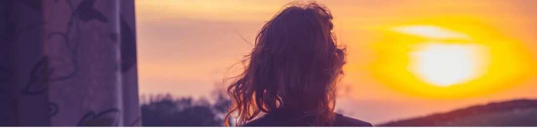 Woman with messy hair looks out the window toward a sunset