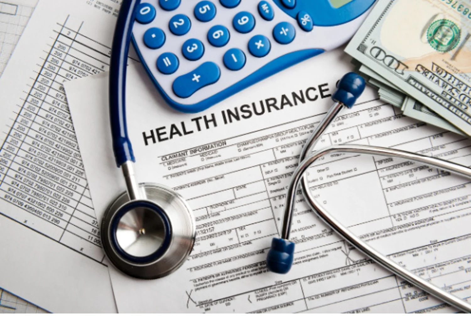 Document that says “HEALTH INSURANCE” covered by stethoscope, calculator, and pile of $100 bills