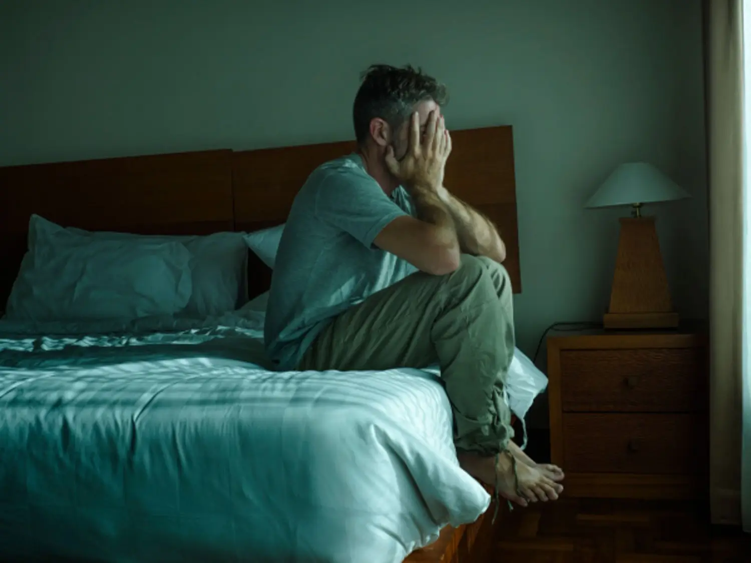 Distraught man going through alcohol withdrawal sits on the edge of his bed holding head in his hands