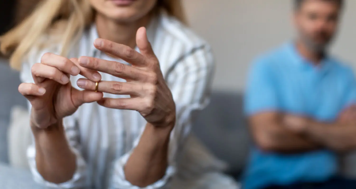 Wife removes wedding ring from finger with upset drug addict husband in background