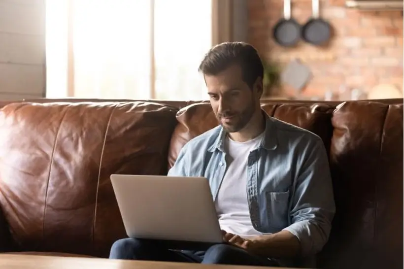Man sitting on brown leather couch researches rehabs on his laptop