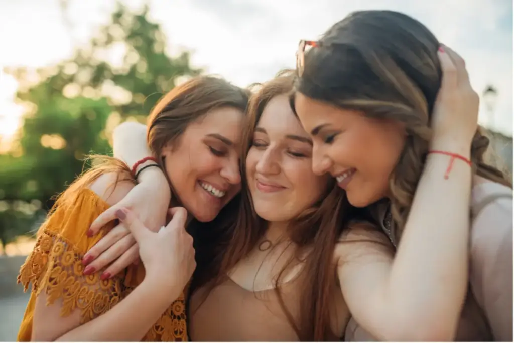 Three young women smiling and embracing outdoors, representing friendship, emotional support, and community during addiction recovery.